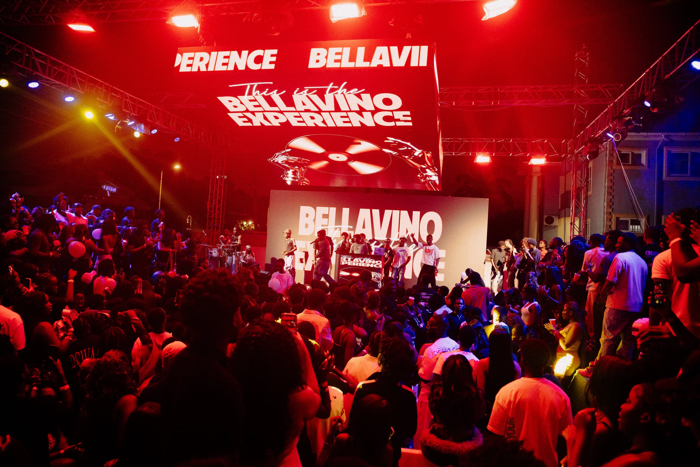 A packed crowd faces the main stage under red lights and two LED screens reading This is the Bellavino Experience